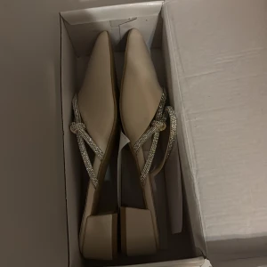 Beige pumps med glittriga remmar - Snygga beige pumps med spetsig tå och låg klack. Skorna har glittriga remmar med strassdetaljer som korsar över foten och ger en elegant touch. Tillverkade i syntetmaterial med en mjuk finish och öppen häl. Perfekta för dig som gillar stilrena och glamorösa skor.