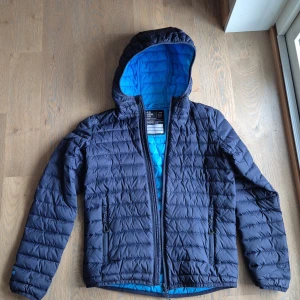 Mörkblå jacka från Neomondo - Snygg mörkblå jacka från Neomondo med huva och quiltad design. Jackan har dragkedja framtill, två sidofickor med dragkedja och coolt blått foder. Perfekt för dig som vill ha en lätt och varm jacka med sportig vibe.