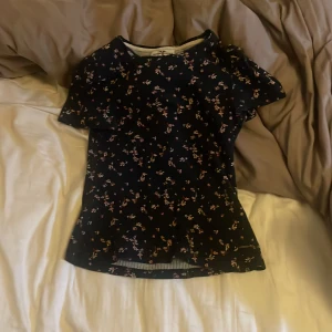 Mörkblå blommig blus med kort ärm - Supersöt svart blus med små blommor i rosa och beige. Blusen har rund hals och korta ärmar, perfekt för dig som gillar romantiska detaljer. Materialet är mjukt och lätt, vilket gör den skön att bära hela dagen. Passar dig som vill ha en feminin och chill stil.