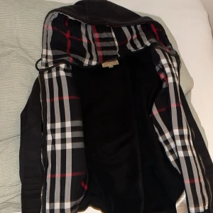 Burberry hoodie med rutor, M - Två snygga hoodiejackor från Burberry i storlek M. Ena är svart med klassiskt rutigt foder i svart, vitt och rött, den andra är grå med beige-rutigt foder. Båda har dragkedja, huva med snörning och mjukt innerfoder. Perfekt för dig som vill ha en ikonisk streetstil. Säljer 1 för 700kr båda för 1400 pris kan diskuteras.