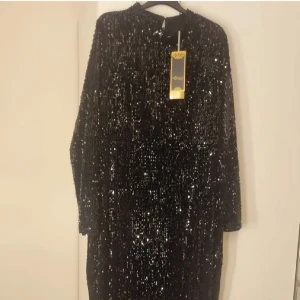 Svart glittrig långklänning Oz Duman - Supersnygg svart långklänning från Oz Duman, täckt med glittrande paljetter som verkligen fångar ljuset. Klänningen har lång ärm och rak passform, perfekt för dig som vill sticka ut. Hög halsringning och elegant siluett.