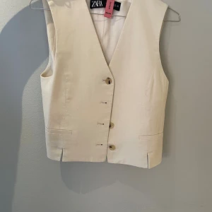 Beige linneväst från Zara - Snygg beige väst från Zara i linne med v-ringad hals och knappar framtill. Västen har två fickor och ett justerbart bälte baktill för perfekt passform. Stilren och enkel design som passar till många olika looks.