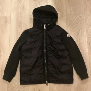 Svart Moncler cardigan - Svart cardigan från Moncler med quiltad framsida, huva och långa ärmar. Jackan har en tydlig Moncler-logga på ärmen och en rejäl dragkedja med Moncler-gravyr. Perfekt för dig som vill ha en clean och stilren look under höst och vinter.