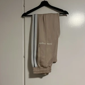 Beige mjukisbyxor från Palm Angels - Snygga beige mjukisbyxor från Palm Angels med vita och svarta ränder längs sidorna. Byxorna har elastiska muddar vid bensluten och Palm Angels-logga tryckt på ena benet. Perfekt för en chill och sportig look.