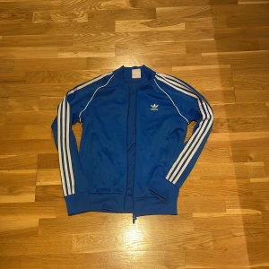 Blå Adidas tröja med dragkedja  - Säljer en klassisk blå Adidas tröja med vita ränder längs ärmarna och Adidas-logga på bröstet. Jackan har dragkedja framtill, ribbade muddar och långärmad design.