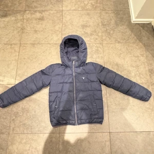 Mörkblå pufferjacka från GANT - Snygg mörkblå pufferjacka från GANT med huva och dragkedja framtill. Jackan har quiltad design, två sidofickor och GANT-logga på bröstet. Perfekt för kalla dagar och har en cool, sportig vibe. Passar dig som gillar klassisk stil med modern touch.