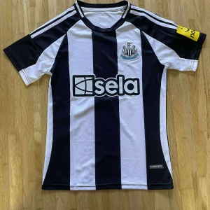 Newcastle United randig fotbollströja - Säljer en officiell Newcastle United fotbollströja från Adidas med klassiska svarta och vita breda ränder, klubbmärke på bröstet och sponsorlogga 'sela' framtill. Kortärmad modell i lätt och ventilerande Aeroready-material. Kaptenens armbindel i gult på ärmen.
