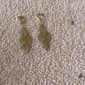 Guldiga clipsörhängen med lövdesign - Snygga clipsörhängen i guld med hängande lövformade detaljer. De har en lätt och luftig design med flera tunna löv som ger ett coolt och naturinspirerat uttryck. Perfekt för dig som gillar statement-smycken med unik form.