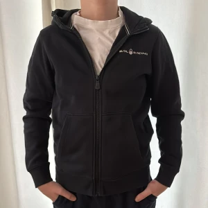Svart hoodie från Sail Racing med huva - Svart hoodie från Sail Racing med dragkedja hela vägen, huva och två framfickor. Diskret logga på bröstet och ett tryck på huvan. Storlek 160. Den har mjuk insida i bomullsmix. Snygg och enkel design som funkar till allt.