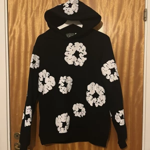 Svart hoodie med vita blommor Denim Tears - Svart hoodie från Denim Tears med stora vita blommor tryckta över hela tröjan, även på huvan. Tröjan har lång ärm, normal passform och är tillverkad i mjuk bomull. Perfekt för dig som gillar streetwear med unik design.pris kan diskuteras.
