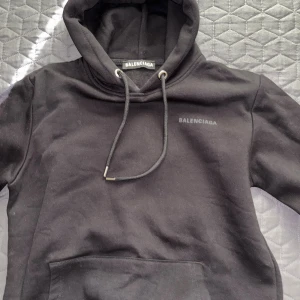Svart hoodie med logga - Svart hoodie med stor logga på ryggen och mindre logga på ryggen. Tröjan har huva med snörning, känguruficka och långa ärmar. Materialet är mjuk bomull och passformen är normal. Storlek S/M