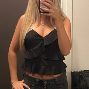 Svart volangig linnetopp - Säljer en svart linnetopp med flera lager volanger och v-ringning. Toppen har axelband och croppad. 