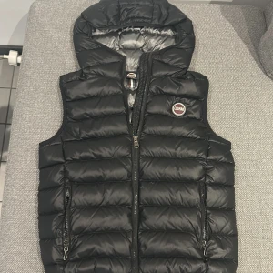 Svart dunväst från Colmar med huva - Snygg svart dunväst från Colmar med quiltad design och huva. Västen har dragkedja framtill, två fickor med dragkedja och Colmar-logga på bröstet. Perfekt för lager-på-lager och chill höst- eller sommar look 👀 