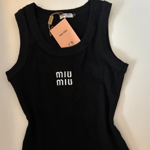 Svart linnetopp från Miu Miu - Supersnygg svart linnetopp från Miu Miu med vit broderad logga framtill. Modellen är croppad och har en tight passform med breda axelband och rund halsringning. Materialet är mjuk bomull som sitter skönt mot kroppen. Perfekt att styla med jeans eller kjol.