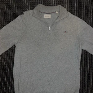 Grå half zip tröja från GANT - Snygg grå tröja från GANT med half zip och liten broderad logga på bröstet. Tröjan har lång ärm och ribbade muddar vid hals och ärmslut. Perfekt för dig som gillar klassisk stil med en modern touch.