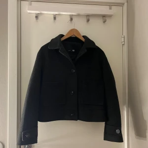 Svart ullrock från 157 - Snygg svart rock från 157 med klassisk krage och stora framfickor. Jackan är croppad och har knappar framtill samt på ärmsluten. Tillverkad i ullmaterial som ger en stilren och varm känsla. 