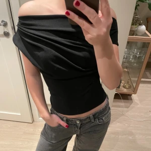 Svart offshoulder topp - Säljer en svart offshoulder topp med snygg drapering över axlarna. Modellen är tight och croppad, perfekt för dig som gillar en stilren och trendig look. Materialet känns mjukt och följsamt mot kroppen. Passar dig som vill ha en cool och modern vibe.