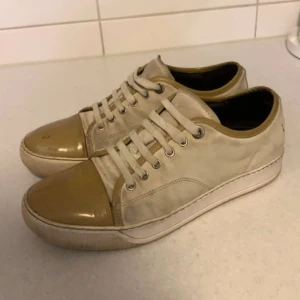 Lanvins - Snygga sneakers i beige med bruna detaljer på tå och runt öppningen. Skorna har rund tå, snörning och platt sula. Ovandelen är i textil och tåpartiet i blankt material som ger en cool kontrast. Perfekt för dig som gillar stilrena och neutrala färger.