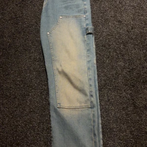 Blå cargopants från Carpe Omnia - Säljer ett par ljusblå cargopants från Carpe Omnia med raka ben och stora fickor framtill. Byxorna har klassisk jeanslook, hällor för bälte och en cool detalj med verktygshållare på sidan. Perfekt för dig som gillar streetwear och funktionella plagg.