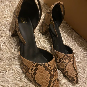 Klackskor  - Snygga pumps från XIT med brunt och svart ormmönster. Skorna har spetsig tå, bred klack och en smal rem runt ankeln. Yttermaterialet har en reptilstruktur som ger en cool och trendig look. Perfekt för dig som gillar att sticka ut med dina skor. Aldrig använt 