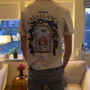 Beige Avenida Casablanca t-shirt - Snygg beige t-shirt från Avenida Casablanca med coolt grafiskt tryck på ryggen och mindre tryck på bröstet. T-shirten har korta ärmar och en relaxed passform, perfekt för dig som gillar streetstyle och unika prints. Materialet känns mjukt och skönt mot huden.