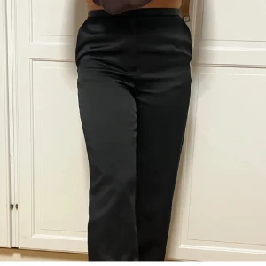 Svarta kostymbyxor med wide fit i siden  - Snygga svarta kostymbyxor med hög midja och wide fit. Byxorna har en slät front, raka ben och en elegant, lite glansig yta som ger en lyxig känsla. Perfekt för dig som gillar stilrena och trendiga plagg med lite extra attityd.