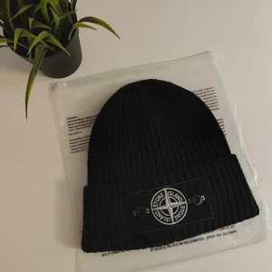 Svart ribbad mössa från Stone Island - Svart ribbstickad mössa från Stone Island med klassisk patch framtill. Mössan har uppvikt kant och är tillverkad i mjukt material som håller dig varm. Perfekt accessoar för att lyfta din streetwear-look.