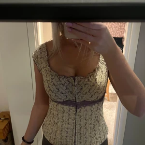 Blommig korsettopp från Charlotte Russe - Supersöt korsettopp från Charlotte Russe i ljusbeige med lila blommönster och spetsdetalj under bysten. Toppen har knappar framtill, volangkant vid axlarna och en feminin, figurnära passform. Perfekt för dig som gillar vintagevibes och romantiska detaljer.