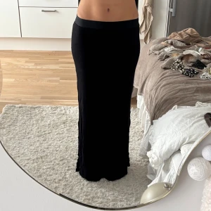 Svart långkjol med slits - Svart långkjol i mjukt och stretchigt material med en snygg slits på sidan. Kjolen har en tight passform och bred resår i midjan för extra komfort. Perfekt för dig som gillar minimalistisk stil och vill ha en enkel men stilren kjol. OBS SISTA BILDERNA