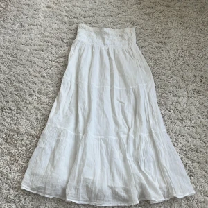 Vit långkjol från Gina Tricot - Säljer en vit långkjol från Gina Tricot med smockad midja och lätt, luftig bomullskvalitet. Kjolen har volangdetaljer och är perfekt för varma dagar. Enkel och stilren design som är lätt att matcha med olika toppar.