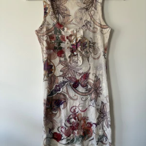 Odd Molly Linne - Supersöt ärmlös Linne från Odd Molly med blommigt mönster i beige, rött, lila och grönt. Linnet har knappar och volangdetalj vid halsen samt ribbad struktur. Perfekt för dig som gillar unika prints och bohemisk stil.