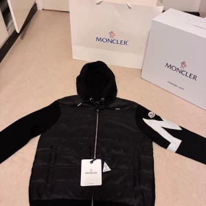 Svart pufferjacka Moncler med huva - Svart pufferjacka från Moncler med huva och snörning. Jackan har stickade ärmar med vit zigzag-mönster och Moncler-logga på vänster ärm. Dragkedja framtill och två sidofickor. Perfekt för dig som vill ha en snygg och unik jacka med streetkänsla.