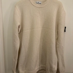 Vit stickad tröja från Stone Island - Säljer en clean vit stickad tröja från Stone Island med klassisk patch på ärmen. Tröjan har rund hals, långärmad design och ett snyggt strukturerat mönster i stickningen. Perfekt för dig som gillar stilrena och exklusiva plagg.