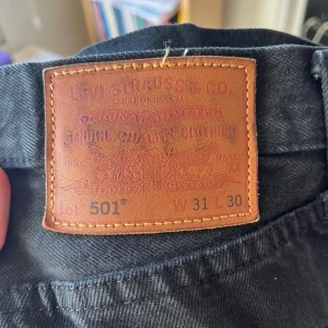 Levi's 501 svarta jeans W31 L30 - Svarta Levi's 501 jeans med klassisk rak passform och fem fickor. Jeansen har knappgylf och är tillverkade i slitstarkt bomullsdenim. Perfekta för dig som gillar tidlös stil och vill ha ett par jeans som funkar till allt.