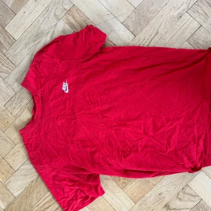 Röd Nike t-shirt i bomull, M - Säljer en röd t-shirt från Nike i storlek M. T-shirten har klassisk passform, korta ärmar och rund hals. Materialet är mjuk bomull och framtill finns en liten vit Nike-logga. Perfekt för dig som gillar sportiga och stilrena plagg.