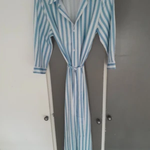 Blåvit randig långklänning med slits - Snygg långklänning med blå och vita vertikala ränder, knappar hela vägen framtill och en markerad slits. Klänningen har lång ärm, krage och ett matchande bälte i midjan för extra stilpoäng. Perfekt för dig som gillar en fräsch och trendig look.