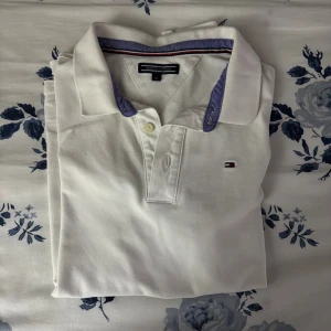 Vit pikétröja Tommy Hilfiger 164 - Vit pikétröja från Tommy Hilfiger med klassisk krage och korta ärmar. Kragen har en snygg blå kant och insida med röd och marinblå rand. Tröjan har två knappar framtill och diskret Hilfiger-logga på bröstet. Perfekt för en clean och fräsch look.