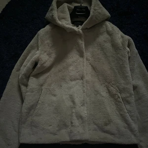 Beige teddyjacka med huva från KappAhl - Mysig teddyjacka i beige från KappAhl, storlek L. Jackan har en stor huva och två sidofickor, passar dig som gillar mjuka material och enkel stil.