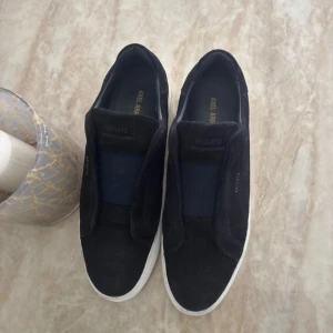 Svarta slip-on sneakers Axel Arigato - Svarta slip-on sneakers från Axel Arigato med vit platt sula och rund tå. Ovandelen är i mocka och insidan har detaljer i skinn. Minimalistisk design med diskret logga på sidan. Perfekt för dig som gillar stilrena och bekväma skor.