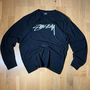 Svart Stüssy longsleave  - Svart långärmad tröja från Stüssy med stort vitt logotryck på bröstet. Tröjan har rund hals och är tillverkad i 100% bomull, vilket gör den skön och lätt att bära. Perfekt för dig som gillar streetwear och vill ha en clean look med tydlig branding.