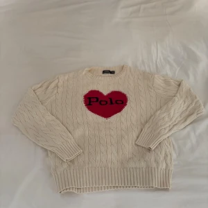 Vit stickad tröja Polo Ralph Lauren - Vit kabelstickad tröja från Polo Ralph Lauren med ett stort rött hjärta och Polo-text på bröstet. Rund halsringning och långa med ribbade muddar. Perfekt för dig som gillar klassisk stil med en twist.