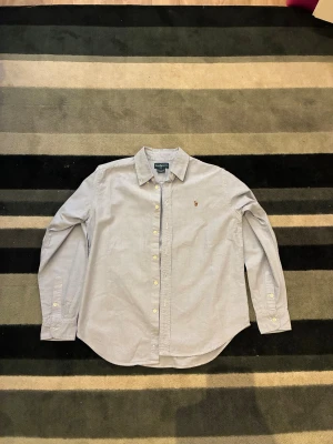 Ralph Lauren Skjorta - Snygg ljusblå skjorta från Ralph Lauren med klassisk krage och broderad logga på bröstet. Skjortan har långa ärmar, vita knappar och är gjord i mjuk bomull. Perfekt för dig som gillar stilrena och tidlösa plagg med premiumkänsla.