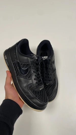 Nike Air Force 1  - Säljer ett par svarta Nike Air Force 1 Low med unik flätad textur på tå och sidor. Skorna har svarta läderpaneler, svart Swoosh i blank finish och klassisk vit yttersula. De har snörning och låg profil, perfekta för dig som gillar stilrena men ändå detaljerade sneakers.