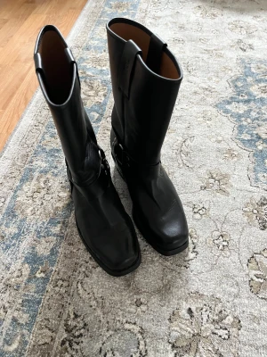 Svarta biker boots från MICC Western - Säljer ett par svarta biker boots från MICC Westerni äkta skinn. Boots med rund tå, grov sula och låg klack. Dekorativa metallringar och nitar vid ankeln ger en cool look. Klassisk modell med högt skaft och dragflikar upptill. Perfekta för dig som gillar edgy stil.