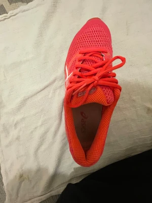 Asics rosa träningssneakers - Snygga Asics sneakers i stark rosa färg med vita detaljer och orange inslag. Skorna har ventilerande meshmaterial, snörning och platt sula med bra grepp. Perfekta för träning och löpning, med sportig design och bekväm passform.