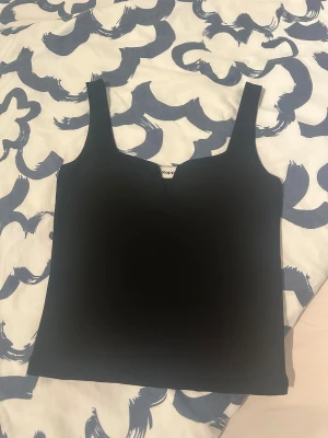 Svart linnetopp från Kappa - Snygg svart linnetopp från Kappahl med breda axelband och en subtil v-ringad detalj framtill. Enkel och stilren design som är lätt att matcha med olika outfits. Materialet känns mjukt och stretchigt, perfekt för dig som gillar minimalistisk stil.