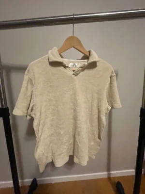 Beige frotté pikétröja - Snygg beige pikétröja från Berns Edition i mjuk frotté. Tröjan har korta ärmar, klassisk krage och relaxed passform. Perfekt för dig som gillar chill och stilren vibe. Passar till både jeans och shorts.