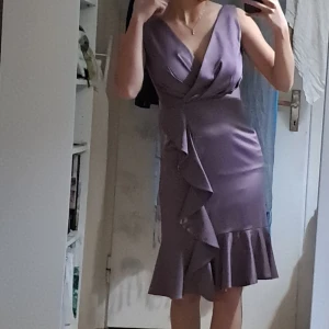 Lila omlottklänning med volang - Säljer en lila omlottklänning med v-ringning och volangdetaljer framtill samt längs fållen. Klänningen är ärmlös och har en figurnära passform med dragkedja i ryggen. Perfekt för dig som vill sticka ut med en elegant och trendig look. Satin, storlek 36