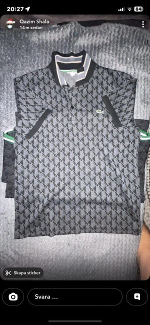 Gråmönstrad henley från Lacoste - Säljer en grå och svart henley från Lacoste med geometriskt mönster och klassisk krage med ränder i svart, grått och vitt. Skjortan har knappar framtill och den ikoniska gröna krokodilloggan på bröstet. Perfekt för dig som gillar stilrena detaljer och snygg design. Helt ny t-shirt