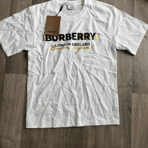 Vit Burberry t-shirt med logga - Vit t-shirt från Burberry med klassisk rund hals och korta ärmar. Stor logotyp i svart och guld på bröstet samt texten 'London England' i svart och guld. Tillverkad i mjuk bomull, perfekt för en clean och stilren look. PS! INGEN TAG FINNS MEN DEN ÄR ALDRIG ANVÄND!!